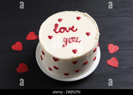 Bento Kuchen mit Text Love You und Papierherzen auf schwarzem Holztisch. St. Valentinstag-Überraschung Stockfoto