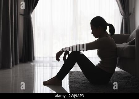 Silhouette einer Person, die an Depressionen im Haus leidet, depressive Frau, die allein auf dem Sofa sitzt, fühlt Stress, traurig und besorgt im dunklen Zimmer Stockfoto
