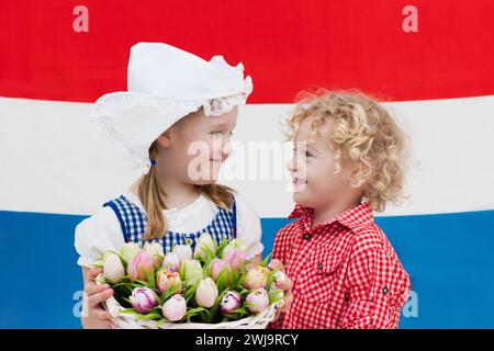 Kleines niederländisches Mädchen und Junge in traditioneller Tracht, Kleid und Hut mit Tulpenkorb an der Flagge der Niederlande. Stockfoto