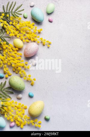 Dieses Bild zeigt ein festliches Osterarrangement aus pastellfarbenen Eiern und hellgelben Mimosa-Blüten auf einer strukturierten hellgrauen Oberfläche. Stockfoto