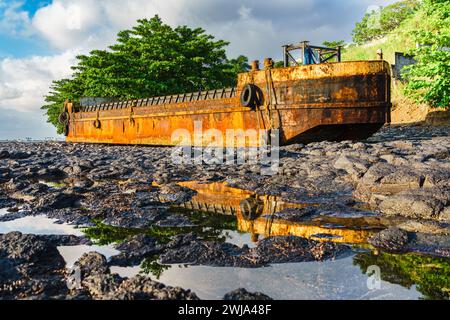 Ein verwittertes, rostiges Schiffswrack befindet sich an der vulkanischen Felsenküste von Praia Emilia in Sao Tome und Principe und spiegelt sich in einem kleinen Wasserbecken unter einem Su Stockfoto