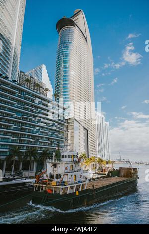 Ein hoch aufragender moderner Wolkenkratzer mit blauer Skyline über dem belebten Frachtschiff in der sonnigen, pulsierenden Stadtlandschaft von Miami, Florida. Stockfoto