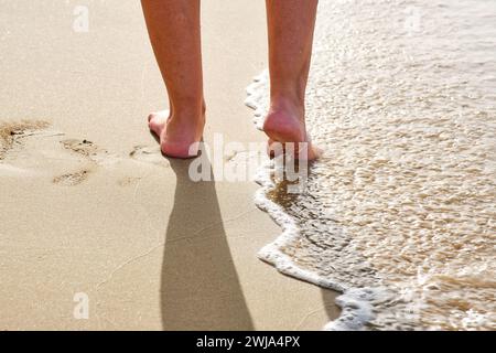 Fußaufnahme einer gesichtslosen Person, die am Sandstrand mit schäumendem Meerwasser in Penon de Ifach in Calpe, Alicante, Spanien spaziert Stockfoto