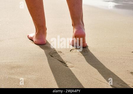 Fußaufnahme einer gesichtslosen Person, die am Sandstrand mit schäumendem Meerwasser in Penon de Ifach in Calpe, Alicante, Spanien spaziert Stockfoto