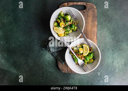 Blick von oben auf eine Schüssel mit gegrilltem Rosenkohl, gewürzt mit Knoblauch und Gewürzen, garniert mit einer Zitronenscheibe, serviert auf einem Holzbrett mit einer Gabel Stockfoto