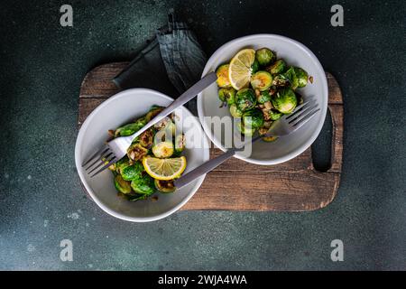 Blick von oben auf eine Schüssel mit gegrilltem Rosenkohl, gewürzt mit Knoblauch und Gewürzen, garniert mit einer Zitronenscheibe, serviert auf einem Holzbrett mit einer Gabel Stockfoto