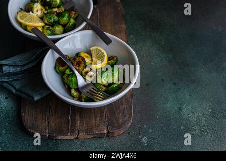 Von oben eine Schüssel mit gegrilltem Rosenkohl, gewürzt mit Knoblauch und Gewürzen, garniert mit einer Zitronenscheibe, serviert auf einem Holzbrett mit einer Gabel Stockfoto