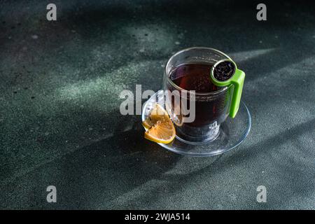 Von oben ein klares Glas Tee mit einer Scheibe Zitrone und einem grünen Teesieb auf einem strukturierten dunklen Hintergrund Stockfoto