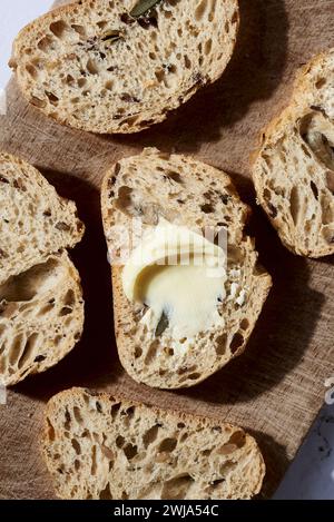 Nahaufnahme frisch gebackener, handwerklich hergestellter Brotscheiben, eine mit einem Klecks Butter, auf einem Holzschneidebrett mit schattigem Hintergrund angeordnet Stockfoto