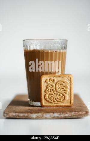 Ein gemütliches Ambiente mit einem klaren Glas warmen Tee Latte, mit einem cremigen Schaum auf der Oberseite, begleitet von einem quadratischen Keks mit einem komplizierten Design, alles wieder Stockfoto