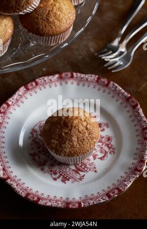 Ein einzelner goldbrauner, mit Zucker bestreuter Muffin sitzt auf einem rot-weißen Teller mit Vintage-Muster, mit einem Kuchenständer und Gabeln im Hintergrund Stockfoto