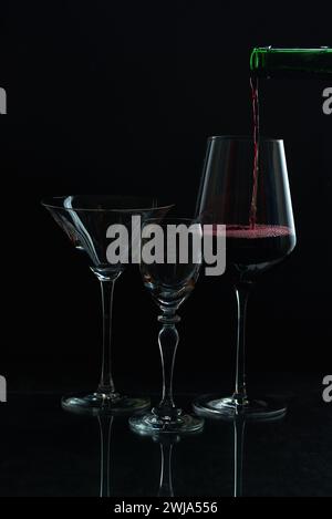 Eleganter Rotwein, der in ein Glas inmitten leerem Stielgeschirr vor dunkler Kulisse gießt Stockfoto