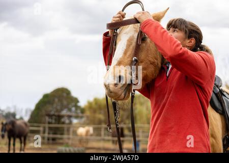 Eine junge Reiterin, die die Zügel und das Zaumzeug auf ihrem braunen Pferd in einem Reitzentrum legt Stockfoto