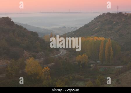 Ein ruhiger Sonnenaufgang bricht über einer malerischen Asphaltstraße inmitten von Herbstlaub auf, während ein sanftes Licht die Landschaft umspült. Stockfoto