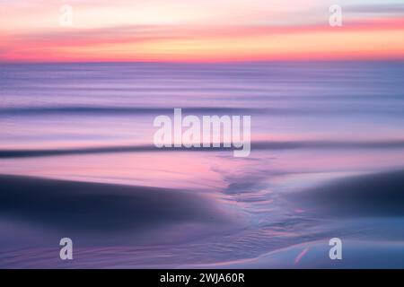 Ätherischer pastellfarbener Sonnenuntergang über ruhigem mediterranem Wasser mit sanften Wellen und sanften Reflexen auf dem Sand Stockfoto