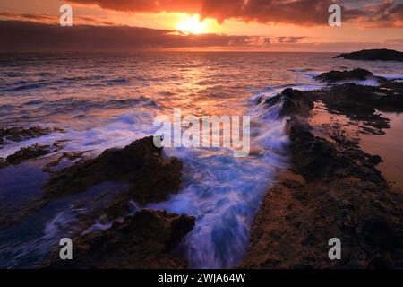 Ein pulsierender Sonnenuntergang über einer felsigen Küste mit dynamischen Meereswellen, die an Land krachen Stockfoto
