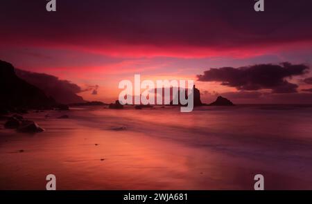 Ein atemberaubender Sonnenuntergang wirft einen violetten Farbton über einem ruhigen Strand, mit Silhouetten von Felsformationen, die vor der sanften Brandung stehen Stockfoto