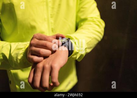 Unerkennbarer junger Mann in gelber Jacke, der die Zeit auf einer intelligenten Armbanduhr überprüft, die am Handgelenk vor dunklem Hintergrund getragen wird Stockfoto