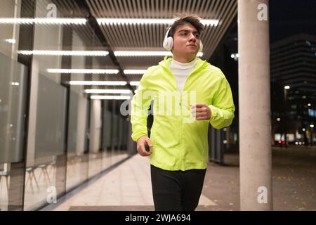 Ernsthafter junger Mann in lässiger Jacke, der Musik in kabellosen Kopfhörern lauscht und wegblickt, während er nachts die Wand eines modernen Gebäudes zusammen läuft Stockfoto
