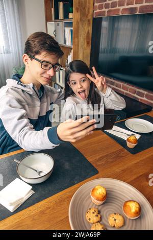 Geschwister machen spielerische Gesichter, während sie ein Selfie mit einem Smartphone am Esstisch während eines Familienwochenendes zu Hause machen Stockfoto