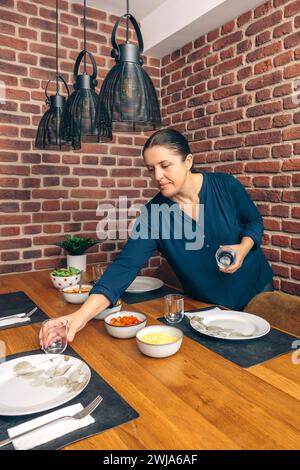 Eine Frau stellt den Esstisch mit Tellern und Gläsern, bereitet sich auf ein Familienessen vor, mit Schüsseln mit Essen auf dem Tisch Stockfoto