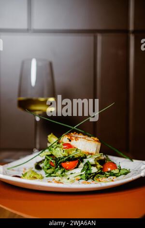 Elegante Präsentation eines Gourmet-Salats mit Kirschtomaten, serviert auf einem stilvollen Teller mit einem Glas Weißwein im Hintergrund Stockfoto