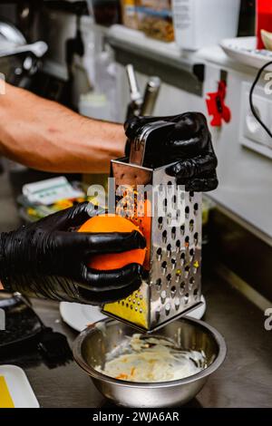 Ein Koch in schwarzen Handschuhen reißt eine Orange über eine Metallschale und bereitet in einer professionellen Küche die Schale für ein kulinarisches Rezept zu Stockfoto