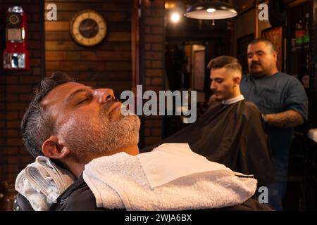 Seitenansicht eines bärtigen Mannes mittleren Alters, der sich auf Stuhlaugen mit Handtüchern um den Hals entspannte, während er auf die Rasur im Friseur gegen Friseur-Givin wartete Stockfoto