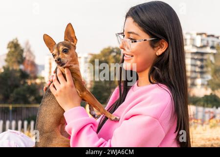 Seitenansicht einer fröhlichen jungen Dame in Brille und rosa Pullover, die den kleinen Chihuahua Hund in der Herbstlandschaft an sonnigen Tagen umarmt Stockfoto