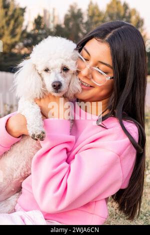 Fröhliche junge Frau in lässigem Outfit und Brille lächelt und umhüllt flauschigen Pudel vor verschwommenen Bäumen auf dem Land Stockfoto