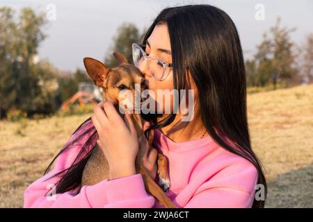 Fröhliche junge Dame in Brille und rosa Pullover umarmt und küsst den kleinen Chihuahua-Hund in der Herbstlandschaft an sonnigen Tagen Stockfoto