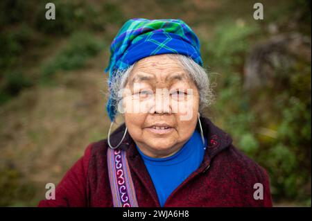 Portrait einer alten Frau in warmer Kleidung mit Riemen und Schal stehend, während sie auf dem Land in Vietnam die Kamera ansieht Stockfoto