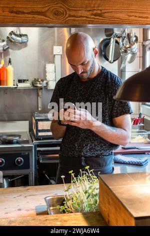 Konzentrierter bärtiger Koch mit schwarzer Schürze, der neben Küchenutensilien steht und während der Arbeit im Restaurant Zermatt in der Schweiz auf dem Handy surft Stockfoto