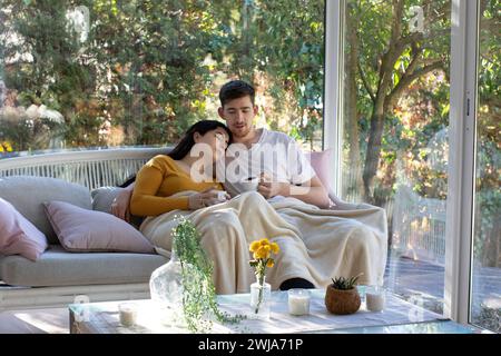 Junger Mann und Frau mit Kaffeetassen auf dem Sofa sitzen und die Beine mit einer Decke bedecken, während sie am Wochenende Zeit zusammen im modernen Wohnzimmer verbringen Stockfoto