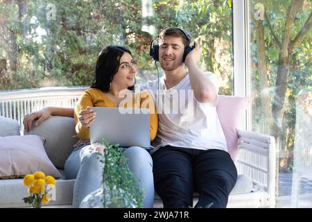 Lächelnde Frau mit Laptop, die jungen bärtigen Mann ansieht, während sie coole Musik mit Kopfhörern hört, während sie am Wochenende Zeit miteinander verbringt Stockfoto