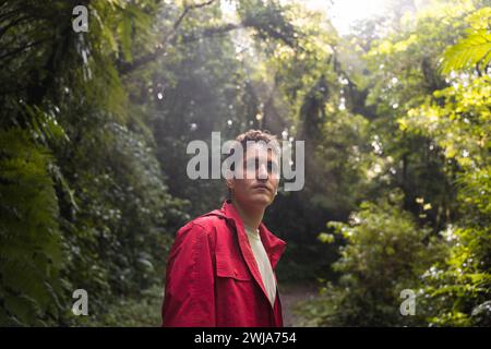 Junger erwachsener Mann in einer roten Jacke, der einen dichten grünen Wald erkundet, voller Staunen. Stockfoto
