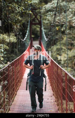 Ein Mann in Wanderbekleidung überquert eine rote Hängebrücke inmitten üppiger Vegetation und fängt das Wesen von Abenteuer und Ruhe ein. Stockfoto