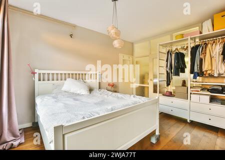 Ein komfortables und warm beleuchtetes Schlafzimmer mit weißem Bett, offenem Kleiderschrank mit Kleidung und bezaubernden Pendelleuchten Stockfoto