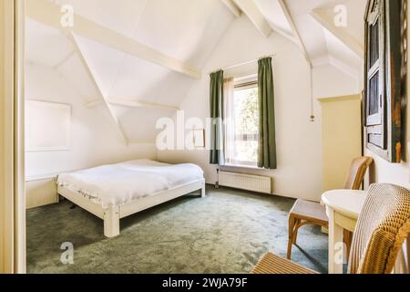 Ein einladendes Schlafzimmer im Dachgeschoss mit einem einfachen Bett, weißen Wänden, freiliegenden Balken, grünen Vorhängen und Blick auf das Grüne vom Fenster. Stockfoto