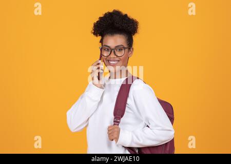 Lächelnde junge schwarze Frau mit Brille und Rucksack, die auf dem Handy spricht Stockfoto