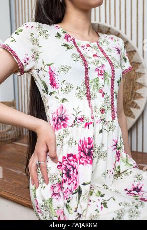 Eine junge Frau posiert in einem Sommerkleid mit Blumenmuster, rosa Blumenknöpfen und komplizierten Spitzendetails. Die entspannte Silhouette und der ausgestellte Rock sorgen für Eleganz Stockfoto