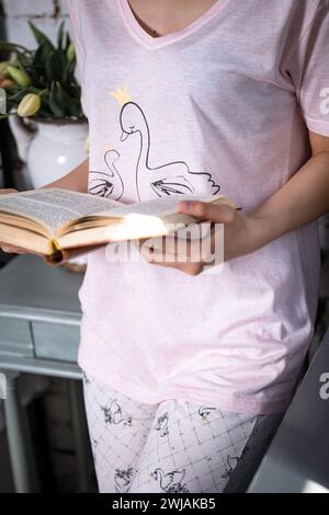 Eine junge Frau mit langen dunklen Haaren in einem rosa Nachthemd, die ein Buch auf einem gemütlichen Bett liest. Sie sieht ruhig und konzentriert aus, umgeben von einem sanften rosa Hintergrund Stockfoto