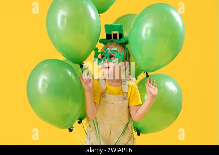 Niedliches kleines Mädchen mit Kobold Hut, Neuheitsbrille und Ballons auf gelbem Hintergrund. St. Patrick's Day-Feier Stockfoto