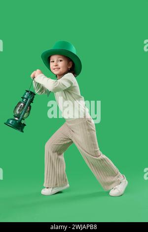 Niedliches kleines Mädchen mit Leprechaun Hut und Laterne auf grünem Hintergrund. St. Patrick's Day-Feier Stockfoto