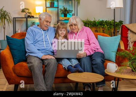 Ältere kaukasische Paare Mann und Frau mit Kind Mädchen Enkelin verbringen Zeit zu Hause zusammen, sitzen auf der Couch im Wohnzimmer mit Laptop Computer. Videos ansehen und am Wochenende Spiele spielen Stockfoto