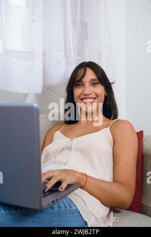 Vertikales Porträt einer jungen brünetten Latina, die zu Hause auf der Couch sitzt und ihren Laptop auf den Beinen arbeitet, lächelt und glücklich in die Kamera blickt. Stockfoto