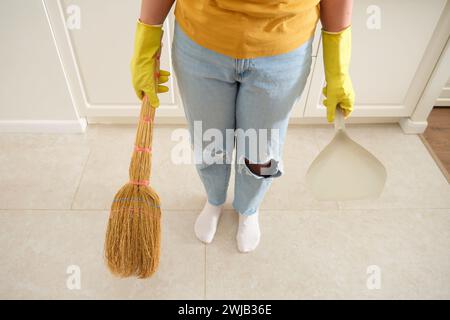 Eine Frau fegt den Boden mit einem Besen und einer Schmutzpfanne in ihrer Küche. Weibliche Hände in gelben Handschuhen beim Reinigen der Küche Stockfoto