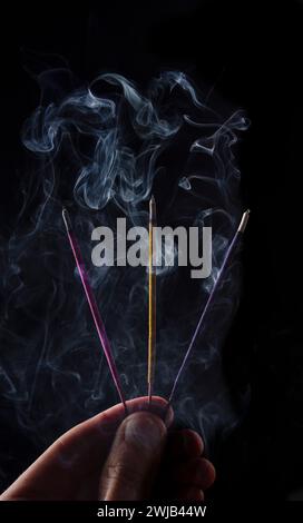 Drei bunte Räucherstäbchen in der Hand eines Mannes. Sticks rauchen auf schwarzem Hintergrund. Stockfoto