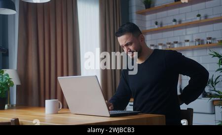 Der Mann, der in der Küche sitzt, erleidet starke Schmerzen in Nacken- und Taillenreibung, um Muskelverspannungen zu lösen, indem er seinen Nacken massiert und eine Pause von der Arbeit macht Stockfoto