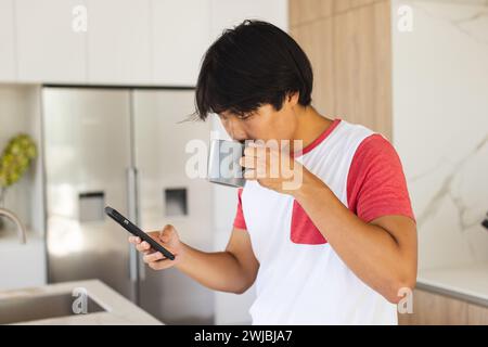 Ein Teenager asiatischer Junge schlürft Kaffee und überprüft sein Handy zu Hause Stockfoto